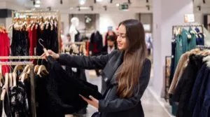 A Woman With Long Hair Carefully Browses Clothes On A Rack In A Clothing Store, Holding A Black Garment That Caught Her Eye. Her Discerning Gaze Mirrors The Precision Of Smoke Damage Restoration, Ensuring Every Detail Is Perfect Before Moving On.