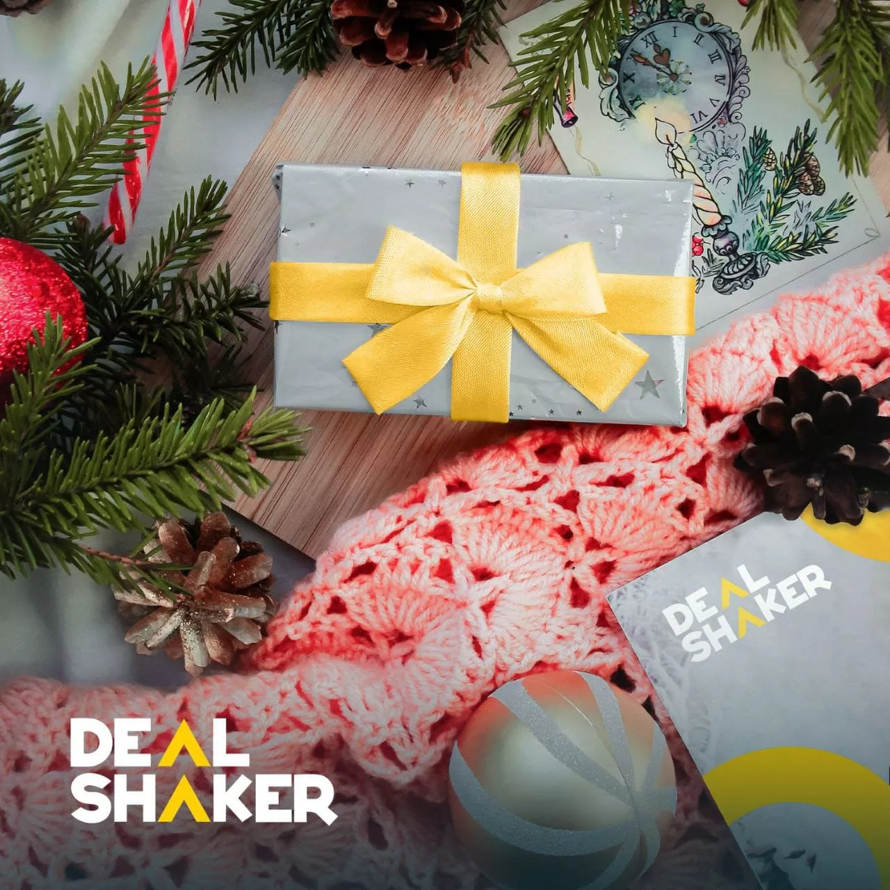 ピンクのかぎ針編みの布の上に、黄色のリボンで包まれたプレゼントが置かれ、松の枝と装飾品に囲まれています。画像の 2 つの角に「DealShaker」のロゴが見えます。