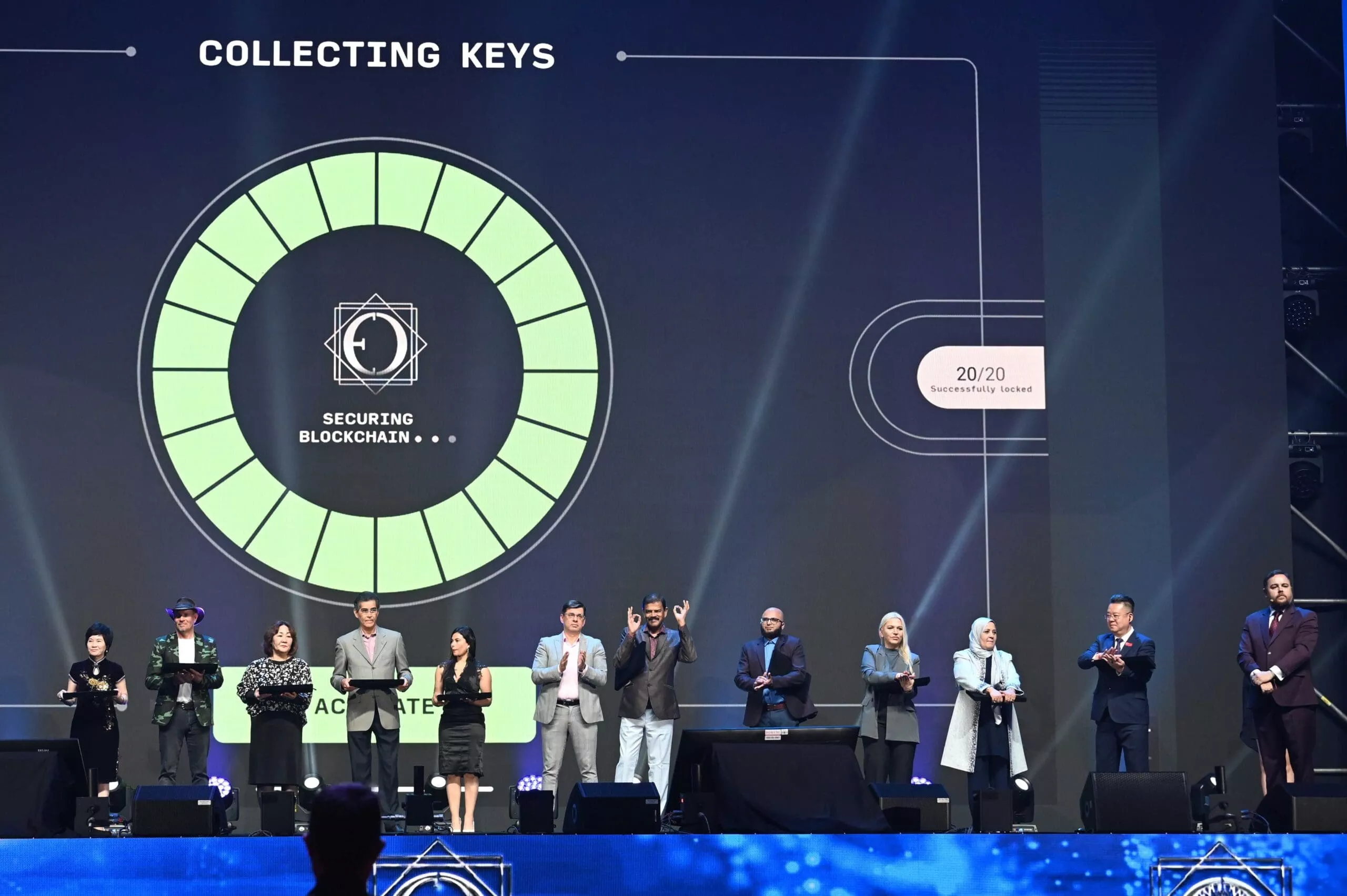 人々ã®ã‚°ãƒ«ãƒ¼ãƒ—ãŒã‚¹ãƒ†ãƒ¼ã‚¸ã«ç«‹ã¡ã€èƒŒå¾Œã®ã‚¹ã‚¯ãƒªãƒ¼ãƒ³ã«ã¯ã€ŒCOLLECTING KEYSã€ã¨ã€ŒSECURING プライベート・ブãƒãƒƒã‚¯ãƒã‚§ãƒ¼ãƒ³ã€ã¨æ›¸ã‹ã‚ŒãŸå††å½¢ã®ã‚°ãƒ©ãƒ•ィックãŒè¡¨ç¤ºã•れã¾ã™ã€‚ç”»é¢ã«ã¯ã€Œ20/20 æ£å¸¸ã«ç™ºè¡Œã•れã¾ã—ãŸã€ã¨è¡¨ç¤ºã•れã€ONE ECOSYSTEM ã¸ã®å–り組ã¿ã‚’強調ã—ã¦ã„ã¾ã™ã€‚