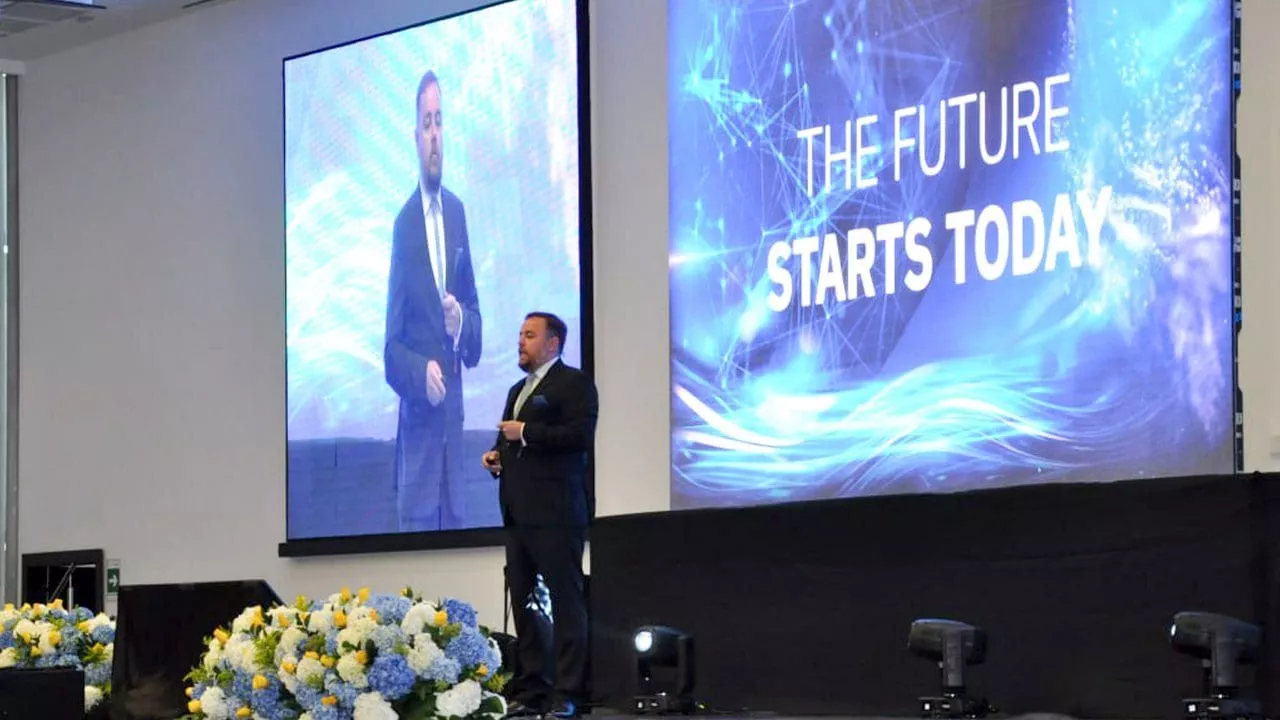 スーツを着た男性が、大きなスクリーンの横のステージに立っています。スクリーンには「THE FUTURE STARTS TODAY」という文字が映し出されています。彼は OES 企業のイベントでプレゼンテーションをしているようです。ステージには花が飾られています。