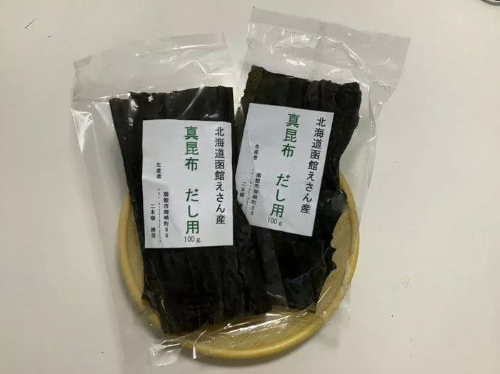 小さな黄色いザルの上に乾燥昆布の密封パックが 2 つあります。日本語でラベルが貼られたパッケージは、道南三大銘柄昆布の最高品質を示しています。