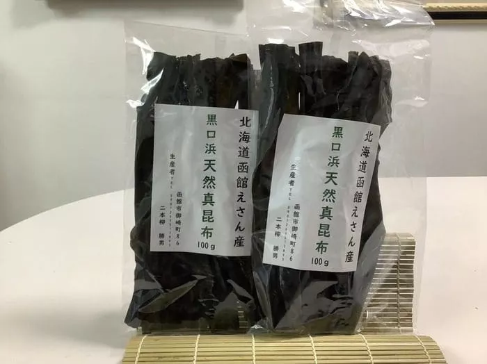 日本語のラベルが貼られた乾燥昆布のパックが 2 つ、竹マットの上に並べられている。 1パックに北海道恵山天然真昆布100gが入っています。最高級の天然昆布を確実にお届けする2袋セットです。