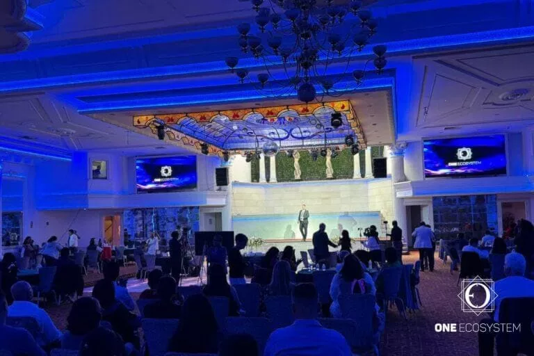 في غرفة المؤتمرات ، يجلس الناس في مواجهة المسرح حيث يقف المتحدث. الغرفة مضاءة بإضاءة زرقاء مع ثريات كبيرة وديكورات سقف. بينما يوضح المتحدث التكنولوجيا المستقلة ، تعرض الشاشة "نظام بيئي واحد".