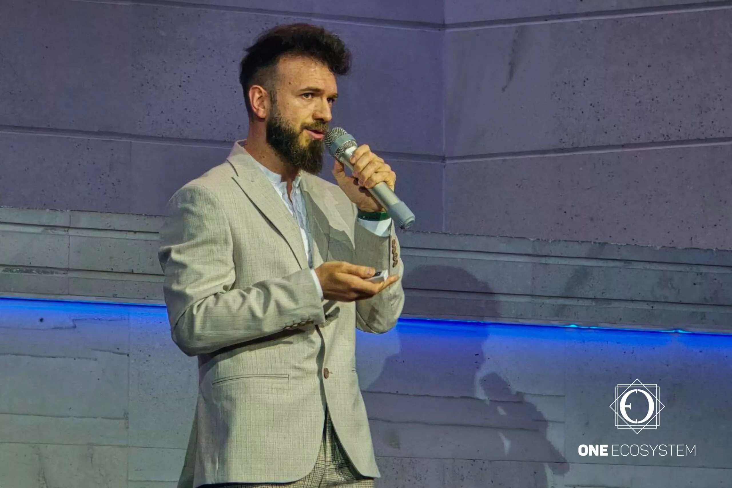 ひげを生やし、髪を整えた男性が、青い線と隅に「ONE ECOSYSTEM」と書かれたロゴがある石の壁を背景に、小さなデバイスを持ちながらマイクに話しかけています。目立つように登場する Ivan Zerkov 氏が、ゲスト購入 と 加盟店 に関連する洞察を提供しています。