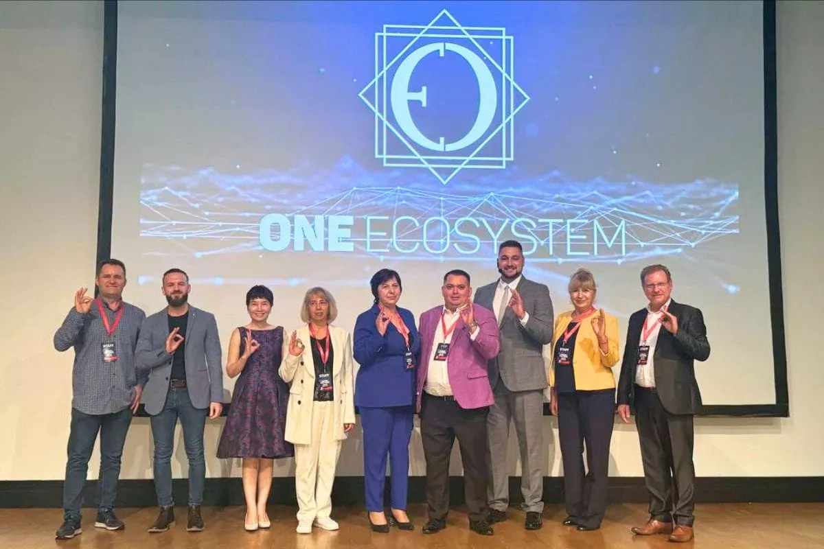 9 人がステージ上に一列に並び、背後で「One Ecosystem」のロゴが光り、それぞれが手振りをしています。このシーンは、エキサイティングな新機能を発表する IMA イベントの一部であり、ルーマニアの革新的精神を世界の観客に伝えています。
