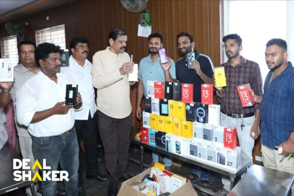 ダバナゲレで開催されたIMAイベントでは、木製の壁と窓のある部屋の中で、一群の人々がさまざまなスマートフォンの箱を並べたテーブルの周りに立っています。