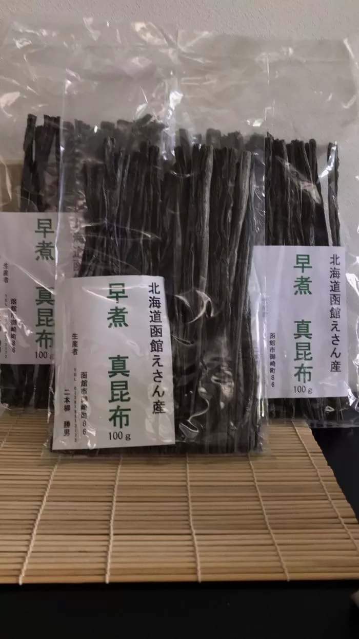 北海道恵山産の乾燥昆布の袋が竹マットの上に立てて並べられ、絶品の早煮昆布の品種を示す日本語のラベルが貼られています。.