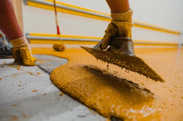 A close-up of a person’s gloved hands spreading yellow epoxy coating onto a concrete floor with a trowel while another person uses a paint roller in the background.