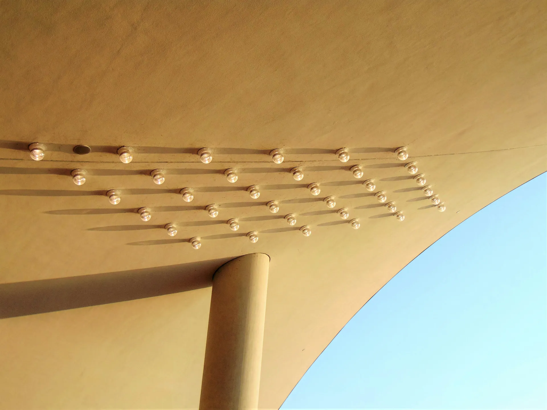 Curved beige ceiling with evenly spaced round lights casting long shadows; a single column supports the structure against a clear blue sky.
