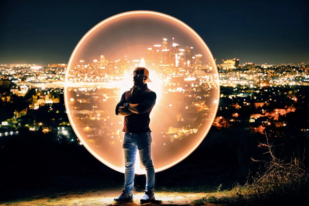 A man stands with arms crossed at night on a hill overlooking a cityscape filled with lights. Behind him, a glowing orange circle creates a halo effect, possibly from light painting. He wears a hat, t-shirt, jeans, and sneakers. The city skyline is blurred in the background.
