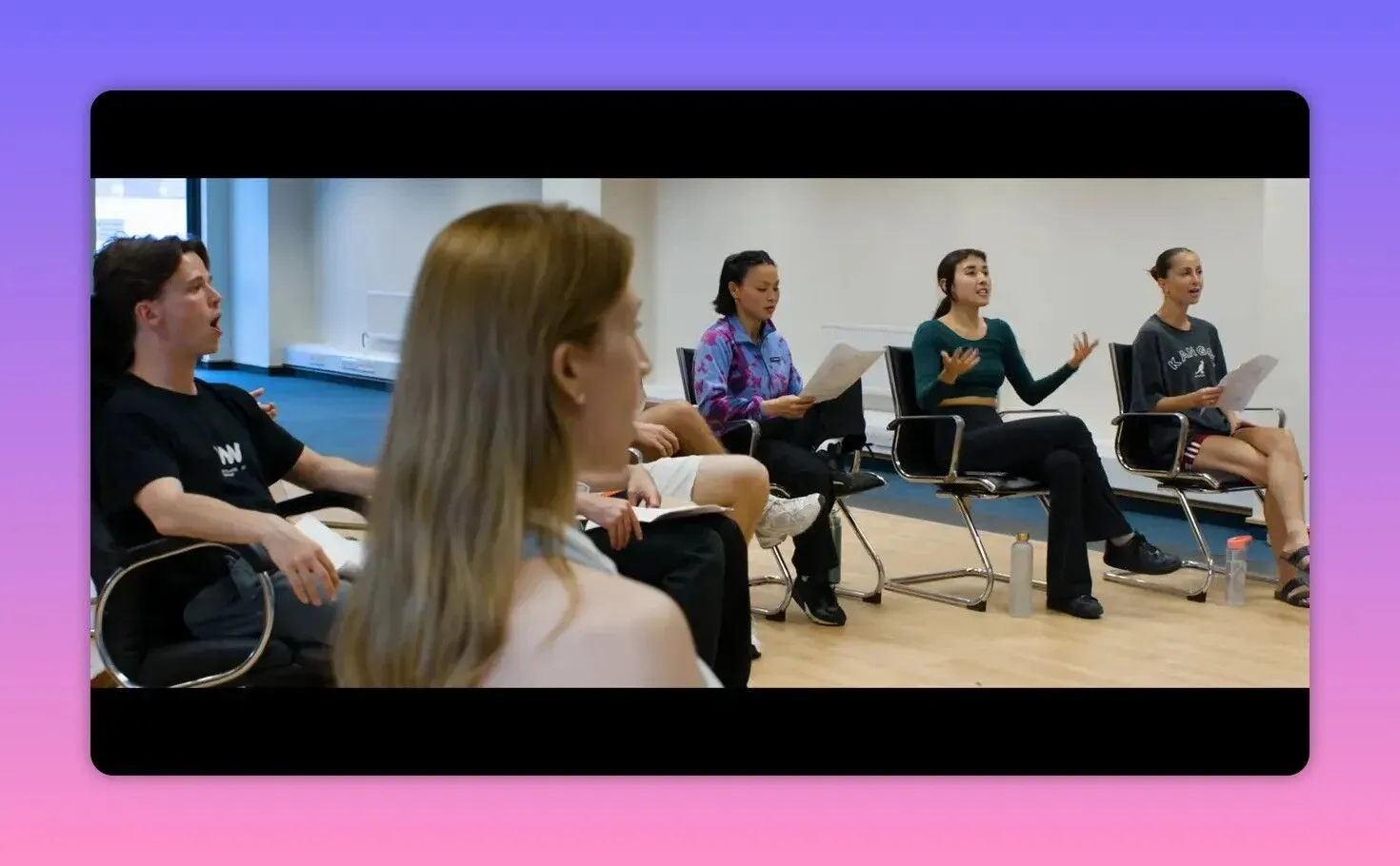 A group of performers seated in a rehearsal room, holding sheets and actively singing and gesturing.