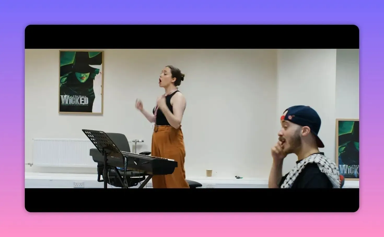 Music trainee conducting and gesturing at a keyboard while a performer listens, with a 'Wicked' poster on the wall.
