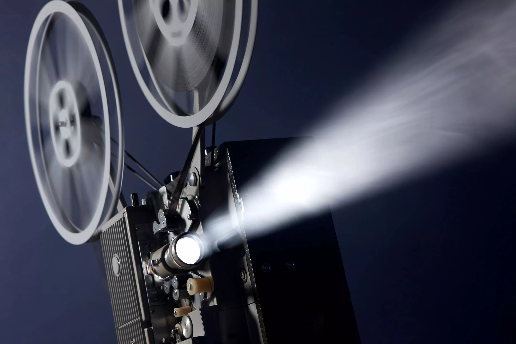 A vintage film projector with two spinning reels projects a bright beam of light forward against a dark blue background, creating a dramatic effect as the film runs through the machine.
