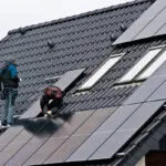 Workers are installing solar panels on a gray metal roof.