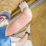 A worker wearing a face mask is installing insulation in an attic.