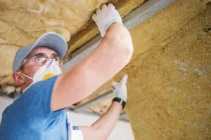 A worker wearing a face mask is installing insulation in an attic.