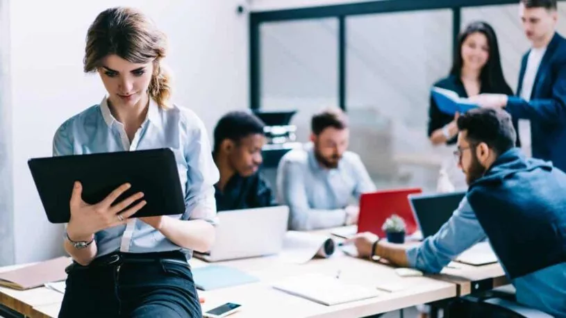 A woman receiving San Marcos tech support on a tablet with coworkers using laptops on a table in the background.