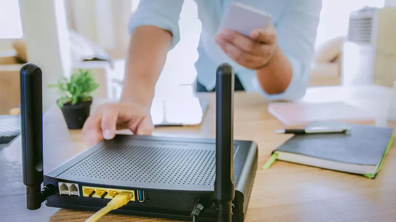 A person adjusts a wireless router on a wooden table, connected with an Ethernet cable. They hold a smartphone in their other hand, with notebooks and a plant visible in the background.
