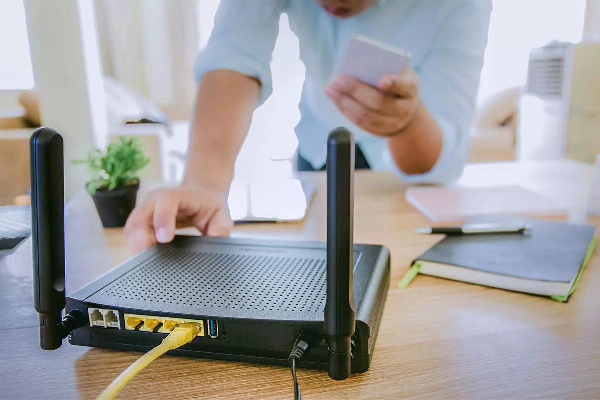A person adjusts a wireless router on a wooden table, connected with an Ethernet cable. They hold a smartphone in their other hand, with notebooks and a plant visible in the background.