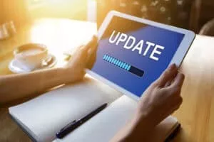 A person holding a tablet displaying a blue screen with the word "UPDATE" and a loading progress bar, with a notebook and a cup of coffee in the background.