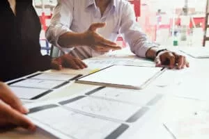 Two individuals are collaborating at a table covered with design mockups for mobile applications, including wireframes and graphs, alongside a tablet displaying a blank screen. One person is gesturing while discussing ideas.