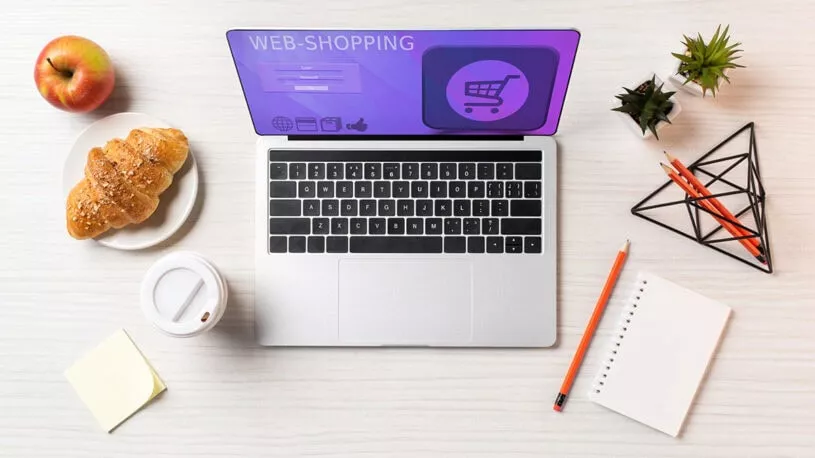 A top-down view of a workspace featuring a laptop displaying a web shopping interface, accompanied by a red apple, a plate with a croissant, a takeaway coffee cup, a sticky note, a blank notepad, and several colored pencils arranged in a geometric holder.