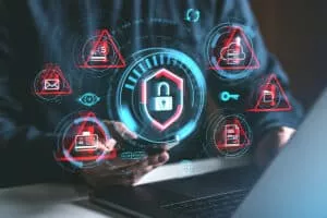 A person holds a smartphone with digital security icons displayed around, including a padlock, alert symbols, and various data protection graphics, set against a blurred laptop.