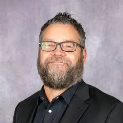 A smiling man with glasses and a full beard is wearing a black blazer over a dark shirt, posing against a textured purple background.