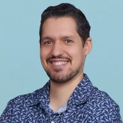 Smiling man with short dark hair and a beard, wearing a patterned blue shirt, against a light blue background.