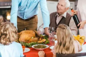 A festive dining table with a roasted turkey at the center, surrounded by various side dishes. A man in a blue shirt serves the turkey while an elderly man in a sweater looks on. Children with curly hair and a girl with long hair sit at the table, watching the scene. A woman pours wine from a bottle.