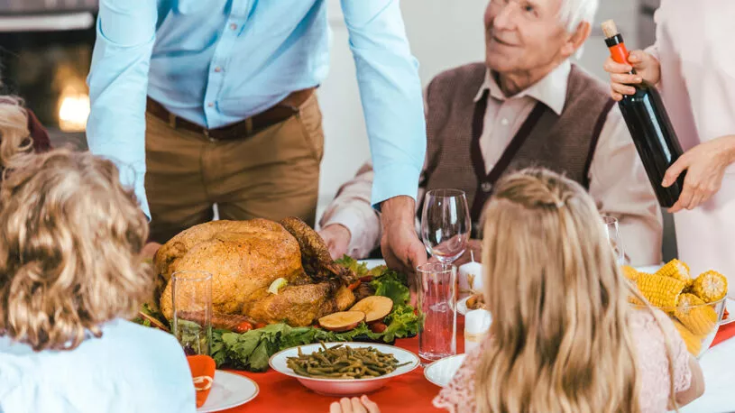 A festive dining table with a roasted turkey at the center, surrounded by various side dishes. A man in a blue shirt serves the turkey while an elderly man in a sweater looks on. Children with curly hair and a girl with long hair sit at the table, watching the scene. A woman pours wine from a bottle.