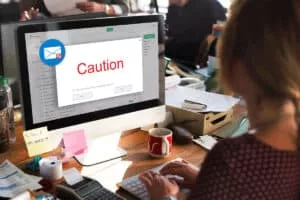 A person is typing on a keyboard in front of a computer displaying an email warning message that says "Caution" in red. The desk is cluttered with paperwork, a red mug, and various office supplies.