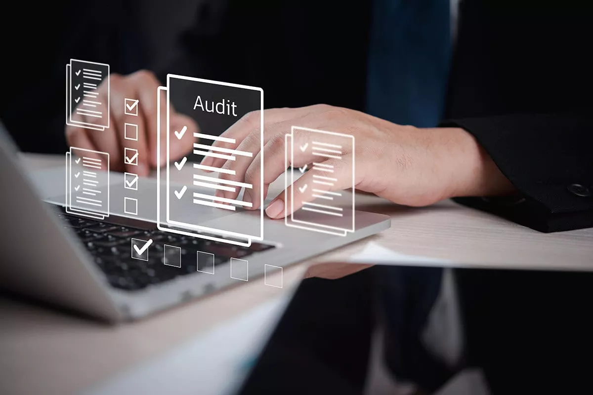 A close-up of hands typing on a laptop with digital audit checklists and documents displayed above the keyboard.
