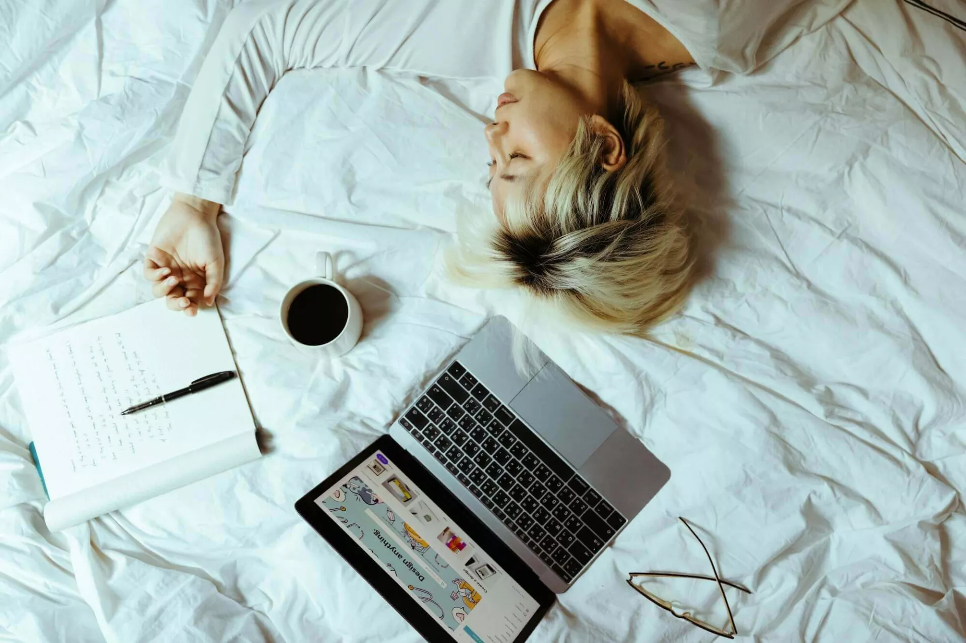 Woman in bed with laptop, notebook, coffee, and glasses. Relatable image for "People Pleaser" blog.
