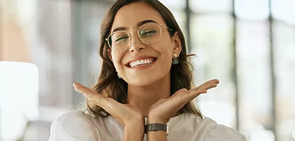 Cheerful business woman with glasses posing with her hands under her face showing her smile in an office. Playful hispanic female entrepreneur looking happy and excited at workplace