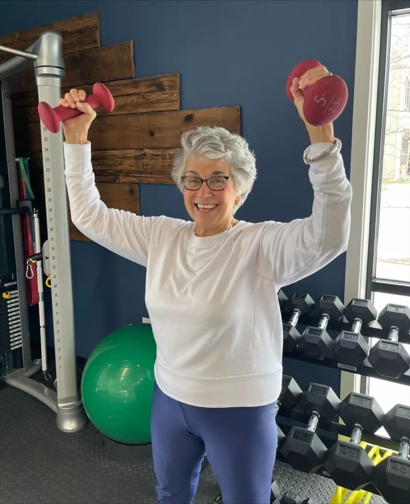 An older woman lifts dumbbells overhead in a gym, surrounded by exercise equipment and a green fitness ball.