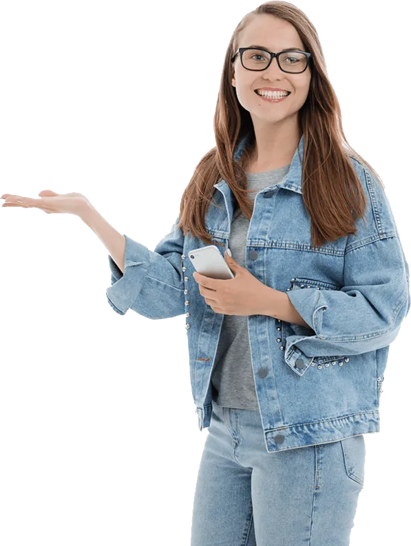 A smiling person with long hair and glasses is standing, holding a smartphone in one hand and gesturing with the other hand. They are wearing a denim jacket and jeans.