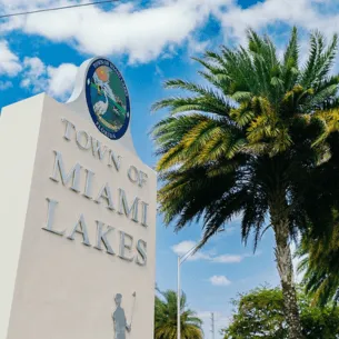 A white monument sign with the emblem of a man playing golf at the top and the text "Town of Miami Lakes" below it. Nearby, a tall palm tree sways under a bright blue sky with scattered clouds, hinting at local businesses like printing services in Hialeah.