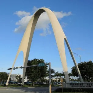 The image shows a distinctive arch-shaped structure in an urban area. The arch is light-colored, soaring into a blue sky with a few clouds. Green trees and a street intersection with a traffic light and a "Sunshine State International Park" sign are visible below, not far from printing service in Hialeah.