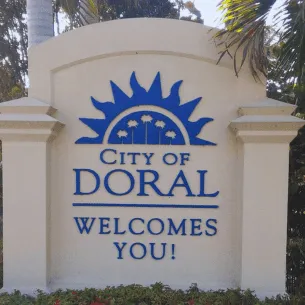 A large beige sign with blue lettering reads "City of Doral Welcomes You!" above a graphic of a sunrise with palm trees. The sign, reminiscent of the quality you’d find at a top printing service in Hialeah, is surrounded by tropical plants and trees in the background.