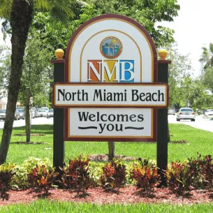 Welcome sign for North Miami Beach, styled with the letters "NMB" at the top, surrounded by landscaped greenery and red flowers. Palm trees and a street with cars are visible in the background on a sunny day, showcasing vibrant scenery you can capture with our printing service in Hialeah.