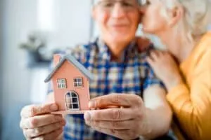 An older man holds a small model house while a woman leans in and kisses him on the cheek, celebrating their decision to use a Reverse Mortgage to Fund Your Retirement.