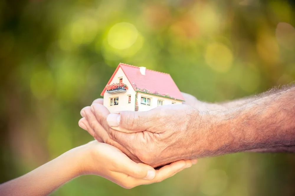 An elderly hand and a young hand hold a small model house together, symbolizing the transfer or sharing of home ownership—an idea echoed by using a Reverse Mortgage to Fund Your Retirement.