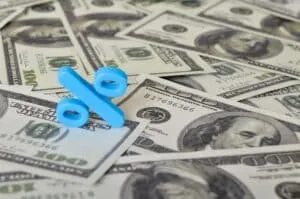 A blue percent symbol sits atop a scattered pile of 100 US dollar bills, visually representing the effects of interest rates on mortgages.