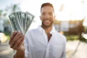 A man in a white shirt stands outdoors, smiling and holding several US $100 bills fanned out toward the camera—perhaps celebrating success after learning about Hard Money vs Traditional Loans.