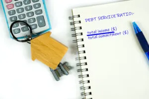 A notebook with "DEBT SERVICE RATIO: total income ($) / total commitment ($)" written on it, a blue pen, calculator, and a house-shaped keychain with keys—essential tools for understanding a DSCR loan.