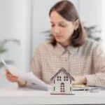 A woman sits at a desk reviewing documents, with a small model house and keys in the foreground—illustrating the careful steps involved in the Bridge Loan Refinancing Timeline.