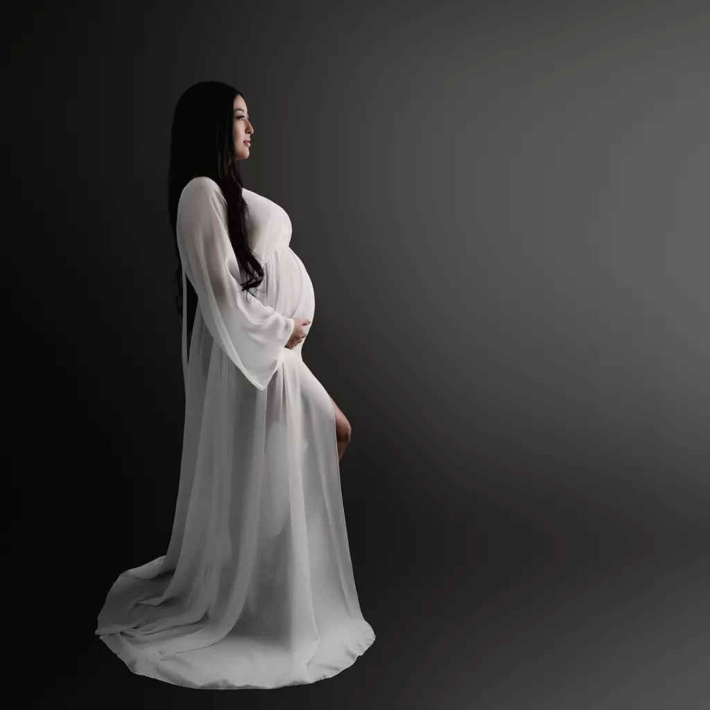 A pregnant woman in a sheer, flowing white gown stands in profile against a dark, gradient background, looking off into the distance.