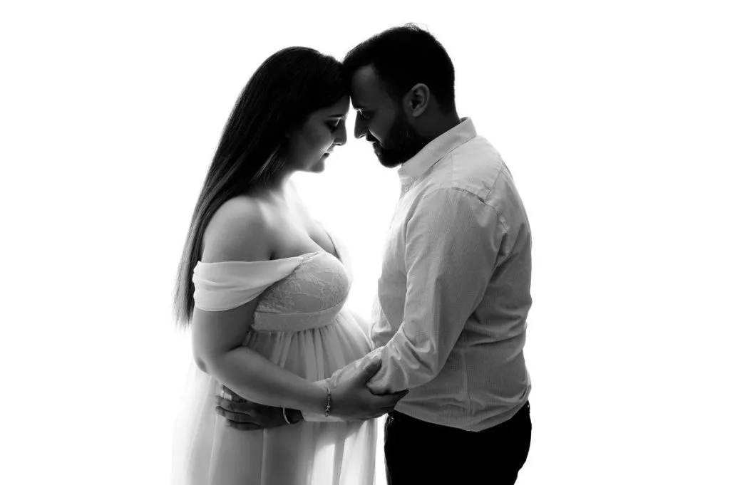 A pregnant woman in a white dress and a man stand facing each other, holding hands and touching foreheads against a white background.