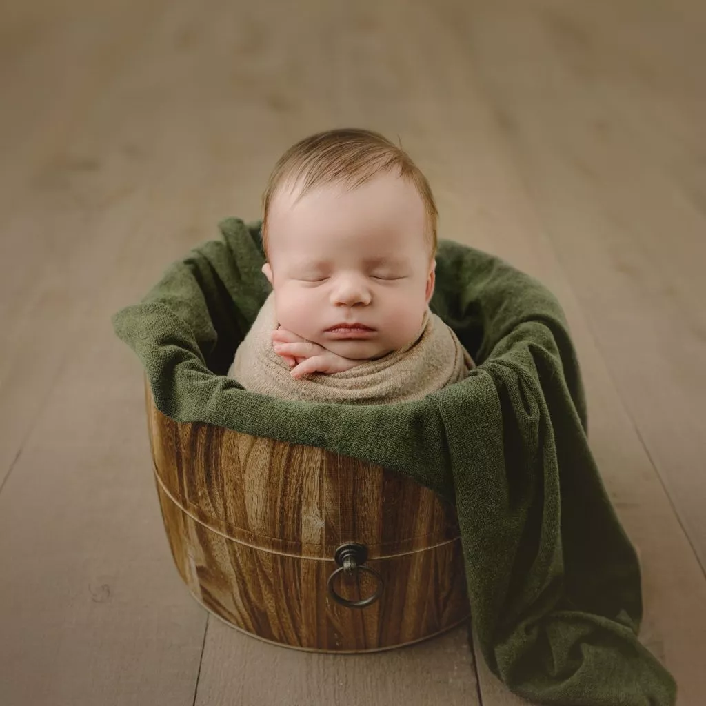A sleeping newborn wrapped in a blanket is posed in a wooden bucket on a wooden floor, with a green cloth draped around them—perfect for capturing timeless newborn photography Vancouver families cherish.